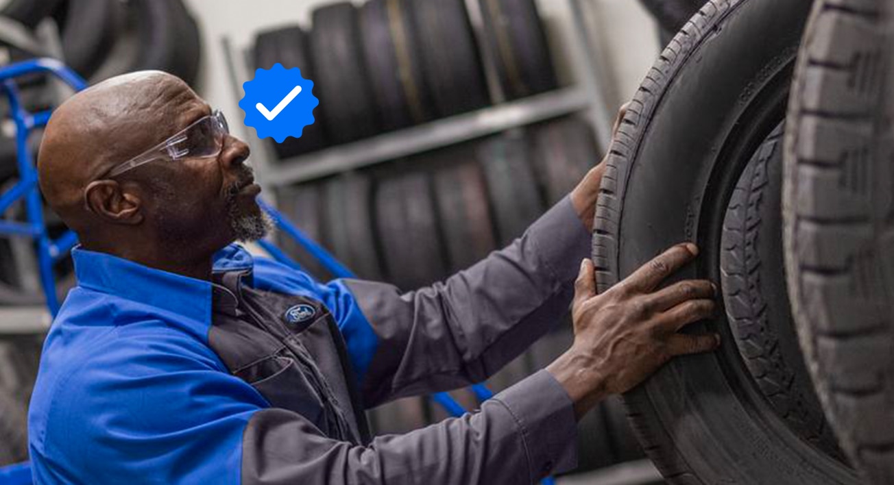 Ford Service Tech holding a changing a tire on a Ford vehicle. A verified badge is supered near the Tech’s head. 