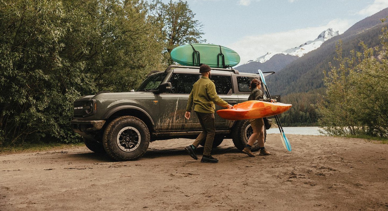 A Ford Bronco is parked by a lake. A couple unloads a kayak and a paddle from the roof rack. There is a mountain in the background.