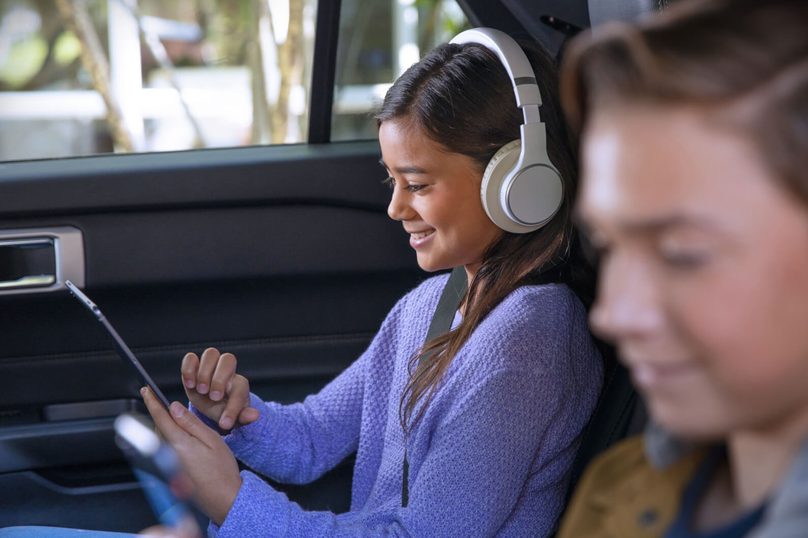 Two children with seatbelts in the back seat of a Ford vehicle looking at their mobile devices and listening to headphones.