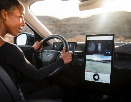 A woman sitting in a Ford Mustang Mach-E with both hands on the steering wheel is looking at a large SYNC 4A screen.