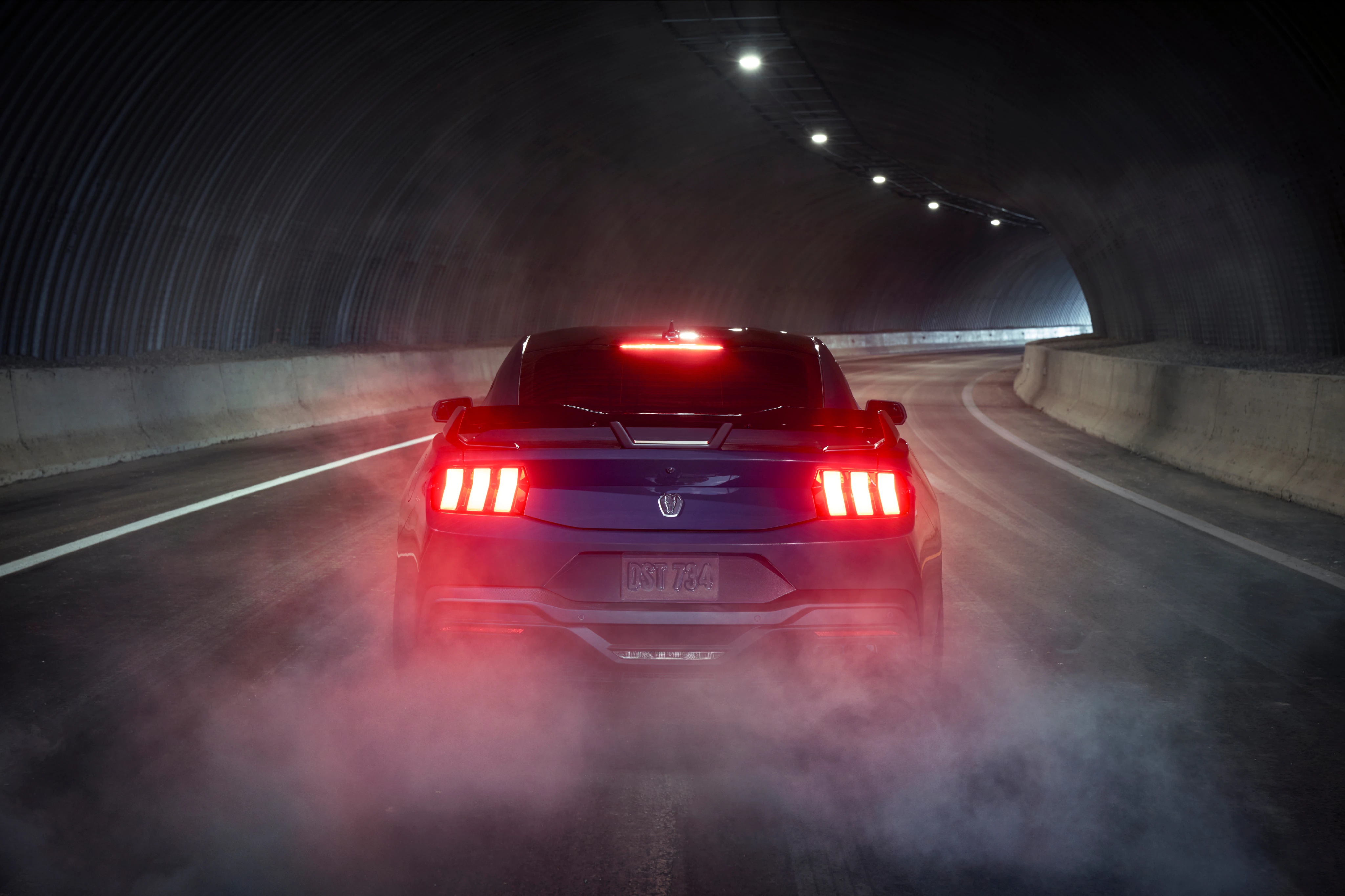 Rear view of a red Ford Mustang Dark Horse driving through a tunnel, illuminated by glowing red taillights and surrounding haze
