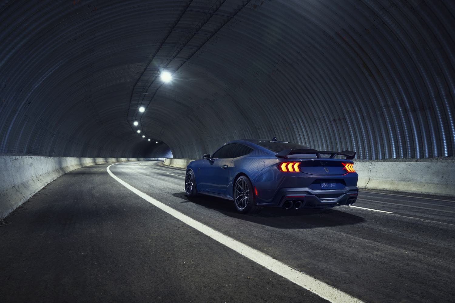 Rear three-quarter view of a dark blue Ford Mustang driving through a curved tunnel, showcasing its taillights and rear spoiler