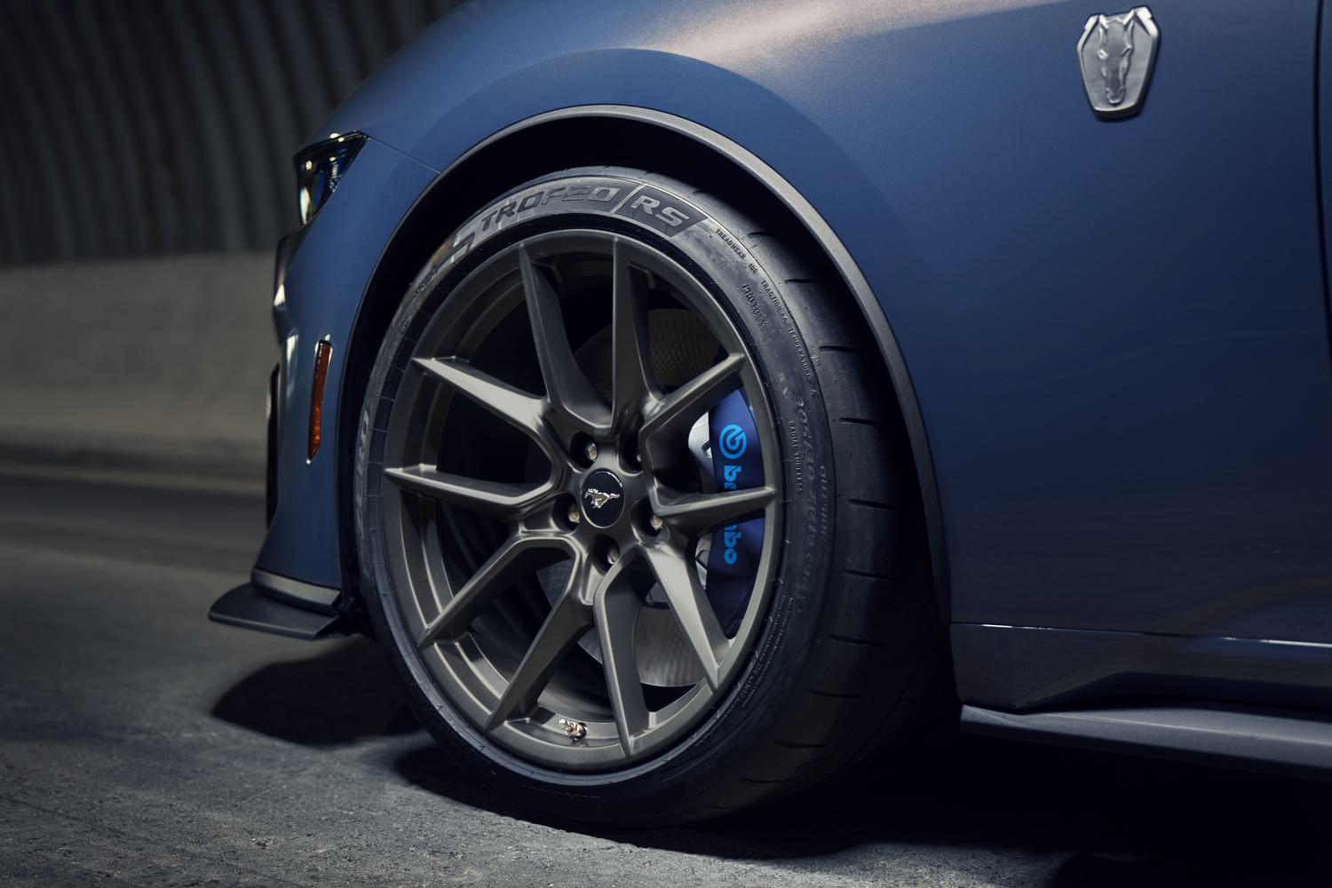 Close-up of a dark blue Ford Mustang's front wheel, alloy rim, and blue Brembo brake caliper with performance tires
