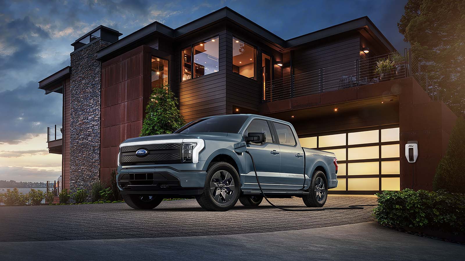 A gray 2023 Ford F-150® Lightning® truck charging outside a home at dusk with an available Ford Accessory charging cable.