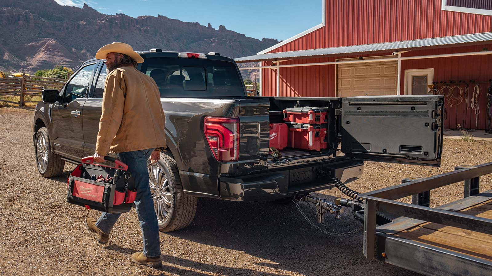 At a ranch near mountains, an F-150® truck owner carries a tool bag away from the open door in the truck’s tailgate.